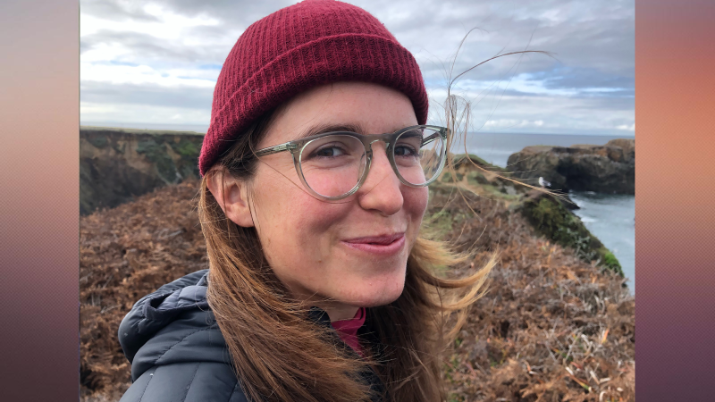 Headshot of Amy Reid outside. She has long hair, glasses, and a red beanie.